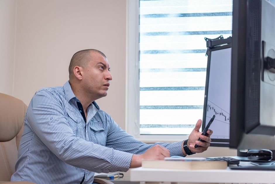 A man intensely studies financial graphs on a computer with a smartphone in hand, working in an office setting.
