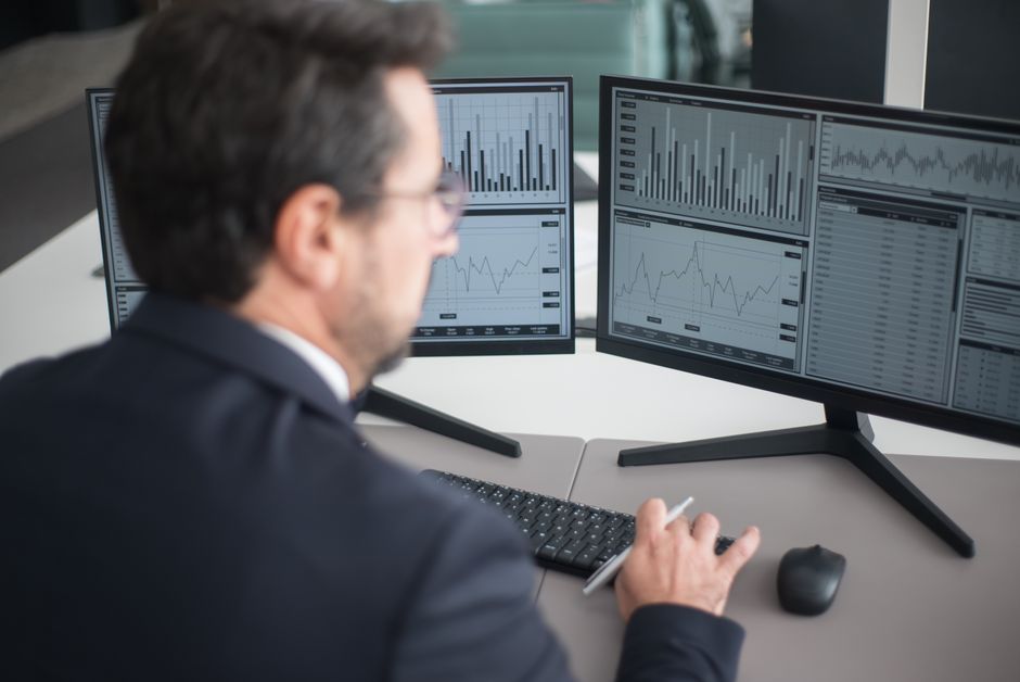 A businessman in a suit analyzing financial charts and graphs on two computer monitors indoors.
